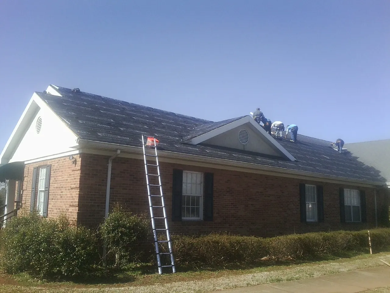Crew tearing shingles off a brick ranch roof with a ladder set — Emergency Roof Repair work by our crew in Rillton