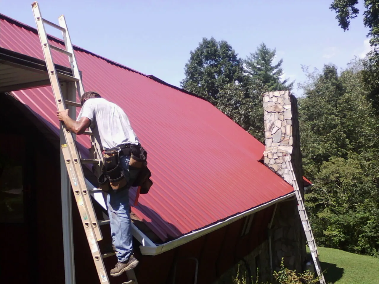 Roofer on a ladder beside a red metal roof and stone chimney — Soffit & Fascia Repair by Roofing Brothers in Rillton